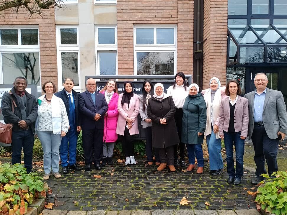 Dreizehn Männer und Frauen stehen nebeneinander in einer Reihe vor einem roten Backsteingebäude. Sie blicken lächelnd in die Kamera. 