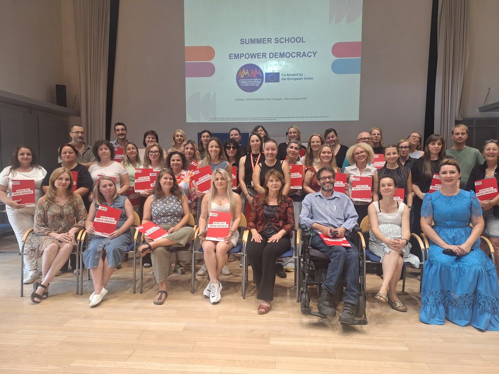Mehrere Menschen befinden sich in einer Aula vor einem angebeamten Bild mit dem Titel "Summer School, Empower Democracy". Die Menschen in der ersten Reihe sitzen auf Stühlen. Alle halten einen roten Umschlag in der Hand.