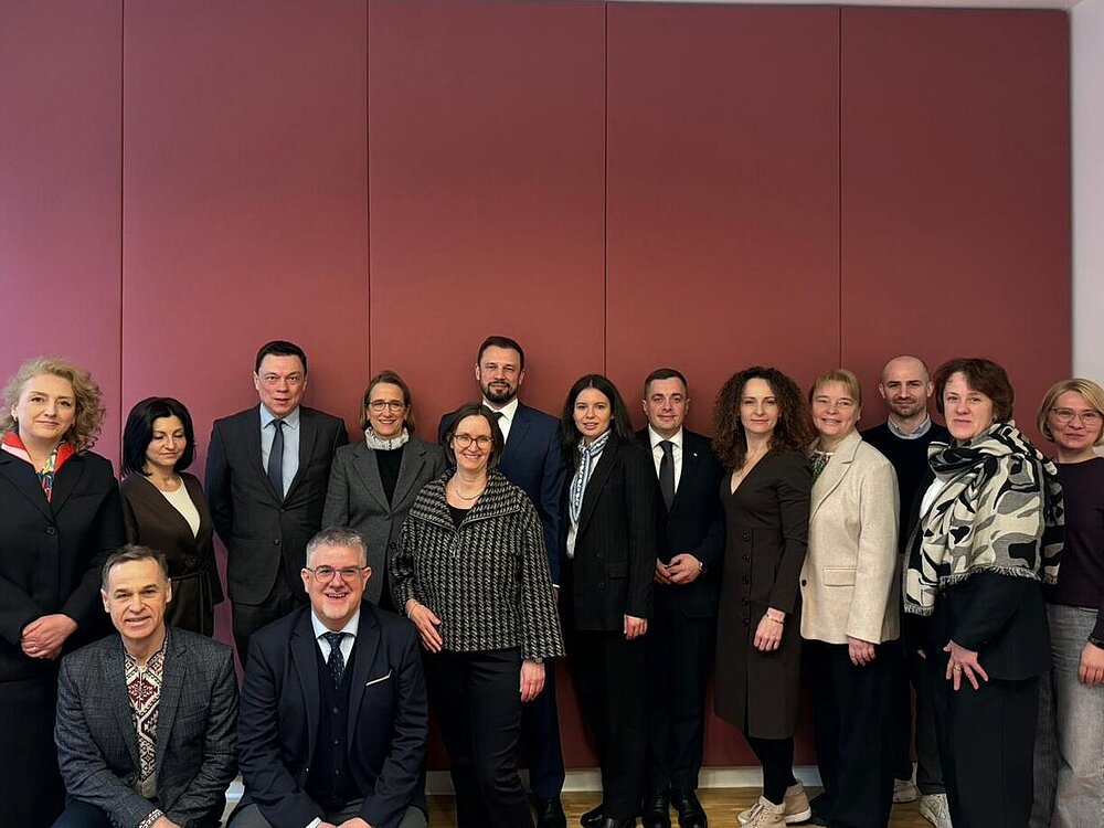 Fünfzehn Frauen und Männer stehen nebeneinander vor einer rötlichen Wand. Sie sind formell gekleidet und blicken in die Kamera.