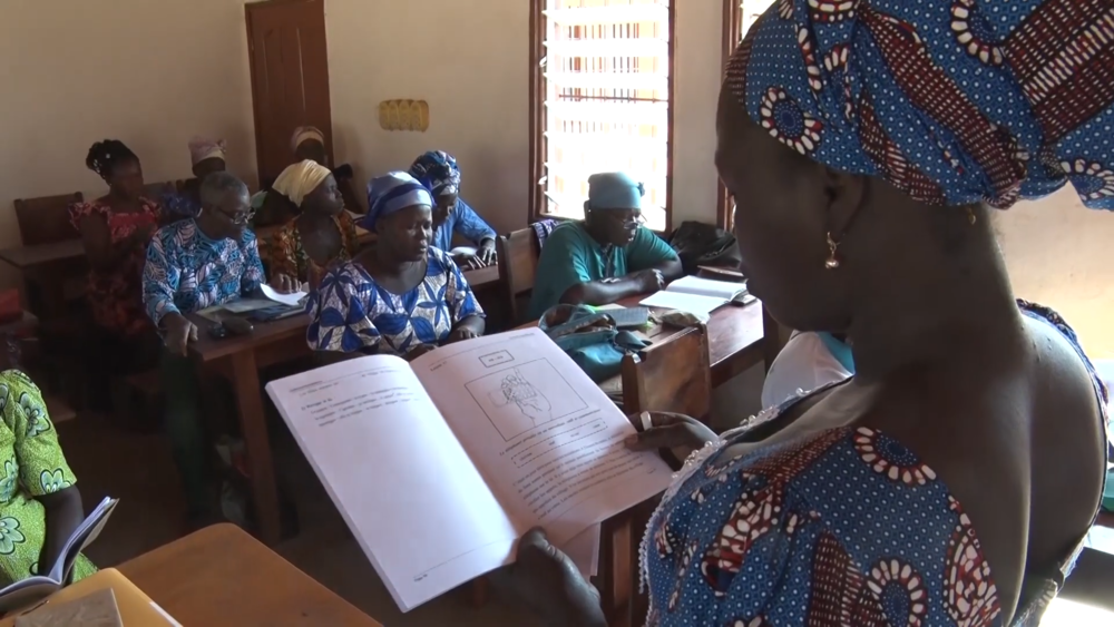 Eine Frau schaut in ein Buch und ist einer Gruppe Erwachsener zugewandt, die in einem Klassenzimmer an Einzeltischen sitzen.
