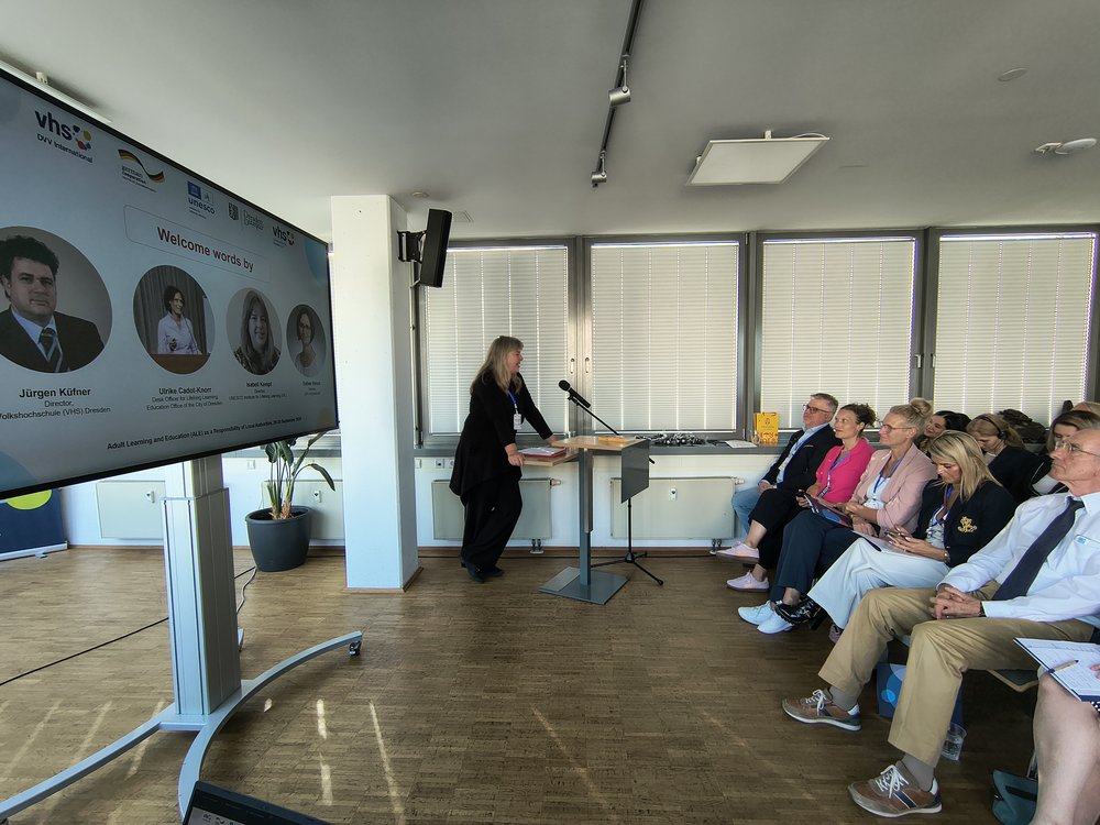 Mehrere Menschen sitzen auf Stühlen vor einem Smartboard. Neben dem Smartboard steht eine Frau an einem Rednerpult und erzählt.