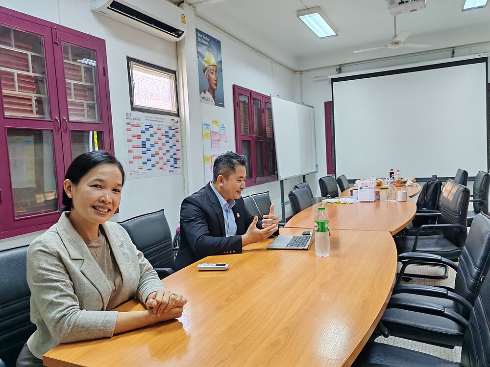 A woman and a man sit at a long conference table. The woman is smiling friendly into the camera. The man is looking at the laptop in front of him and gesticulating. There is a white board at the end of the room and plans, calendars and posters hang on the walls. 