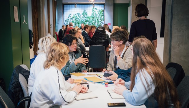 Mehrere Personen sitzen an einem Tisch in einer lebhaften Gruppendiskussion, sie haben Notizbücher, Stifte und Unterlagen vor sich liegen. Im Hintergrund befinden sich weitere Gruppen, die ebenfalls an Diskussionen teilnehmen. 